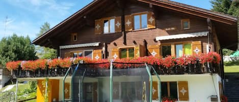 Une maison en bois à deux étages, dotée d’un balcon orné de fleurs rouges, d’un trampoline dans le jardin et, en arrière-plan, d’un ciel dégagé.