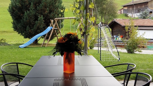Una mesa con un florero, un columpio y un tobogán en un espacio al aire libre con césped y casas al fondo.