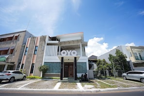 Un edificio de varios pisos con un llamativo letrero 'OYO', un acceso con puerta de seguridad y un estacionamiento con autos y una motocicleta.