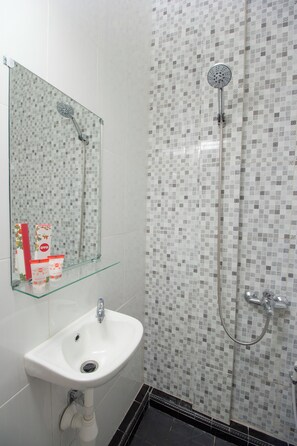 A bathroom with a white sink, a glass shelf, a shower with a handheld showerhead, and a tiled wall. - Irooms (Ngaglik)