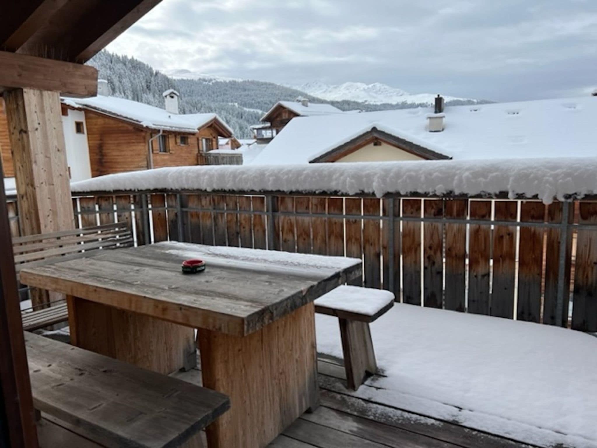 Eine Holzterrasse mit Tisch und Bank, umgeben von einem Holzzaun, mit Blick auf eine verschneite Landschaft mit Häusern und Bergen.