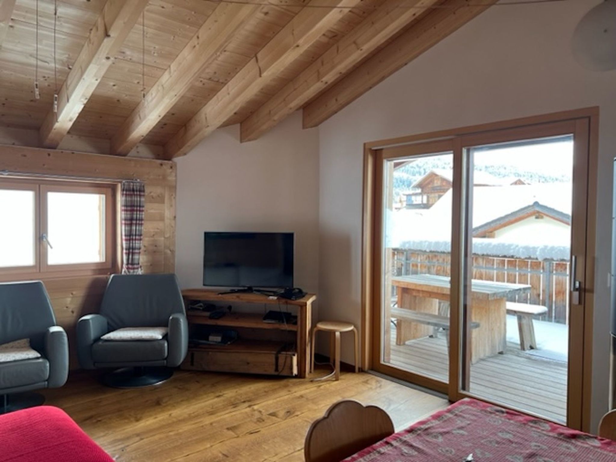 Una habitación con techo de madera, un televisor sobre un mueble de madera, dos sillas, una mesa con mantel rojo y una puerta corrediza de vidrio que da a una terraza exterior.