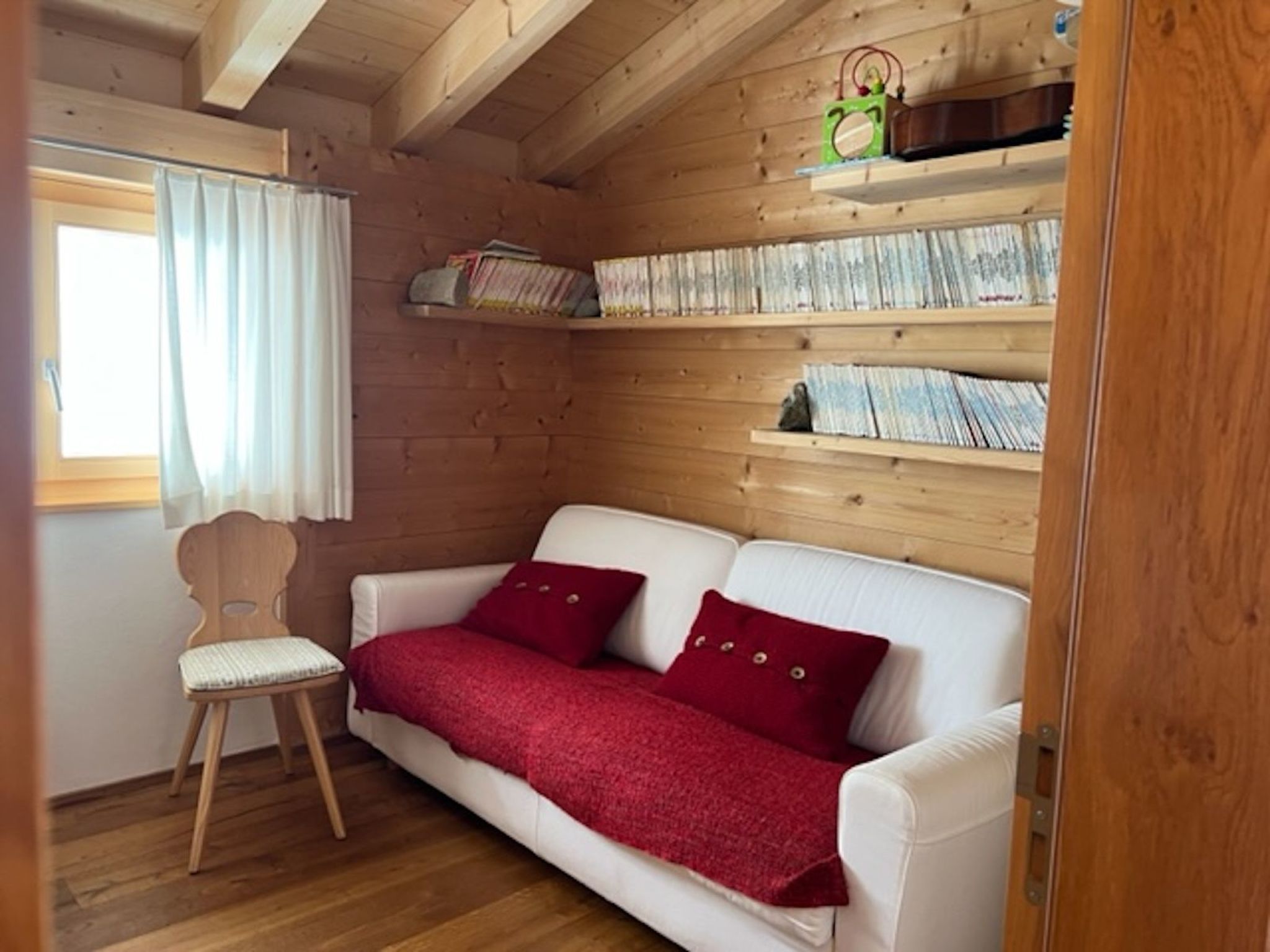 A cozy room with a white sofa, red cushions, a wooden chair, and a bookshelf with books.