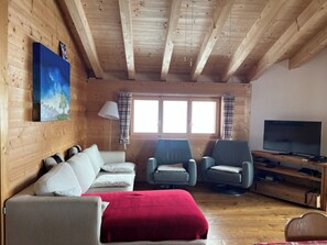 Ein gemütliches Zimmer mit Holzwänden, einer Couch, zwei Sesseln, einem Fernseher und einem Fenster mit Vorhängen.