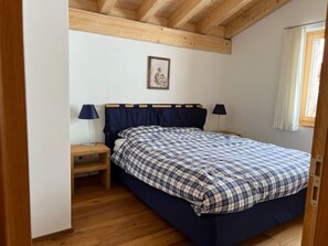 Ein Schlafzimmer mit einem Bett, zwei Nachttischen, einem kleinen Tisch, einer Holzbalkendecke und einem Fenster mit Vorhängen.