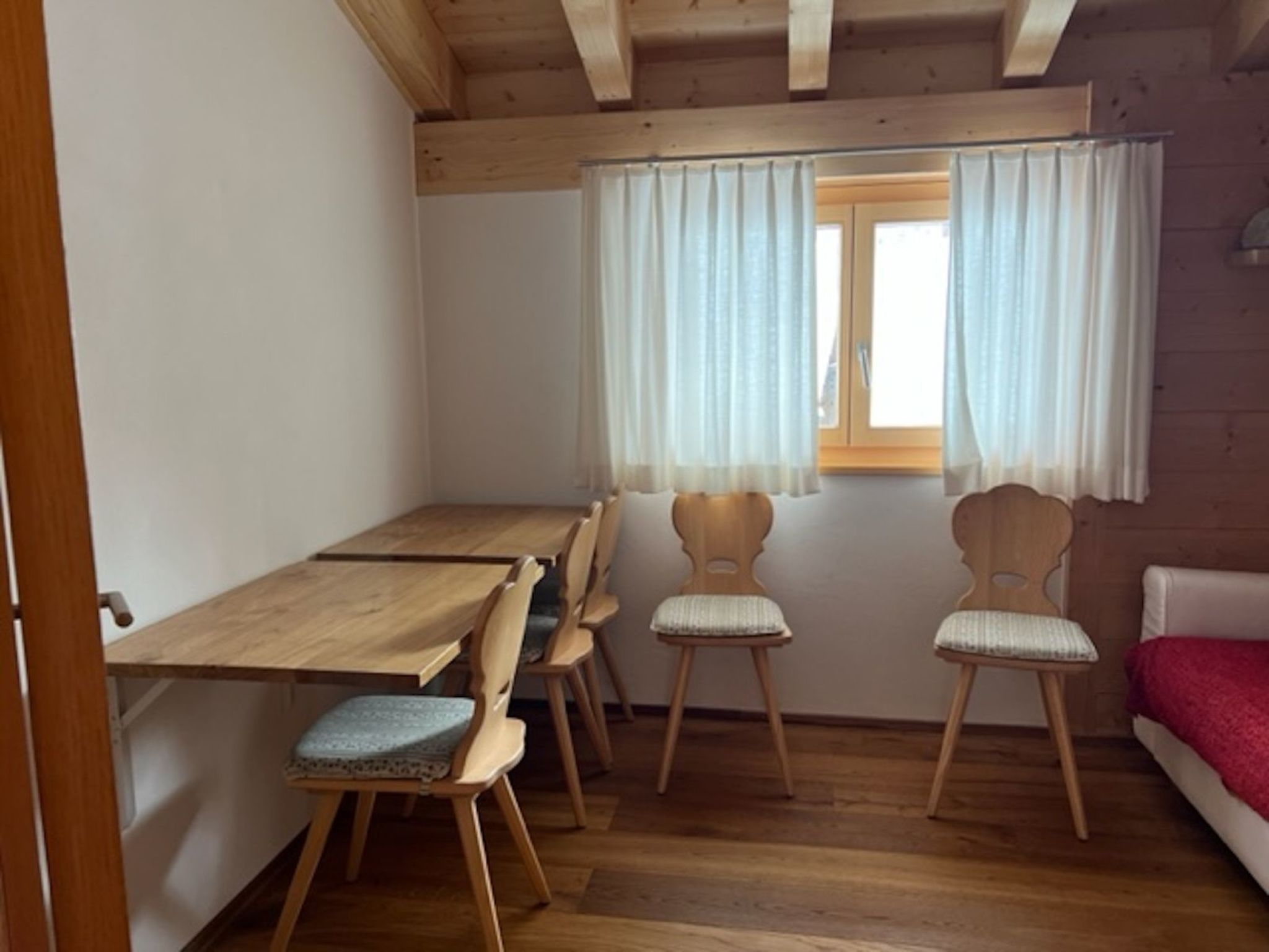 A small room with a wooden table and four chairs, a window with curtains, and a bed with a red blanket.