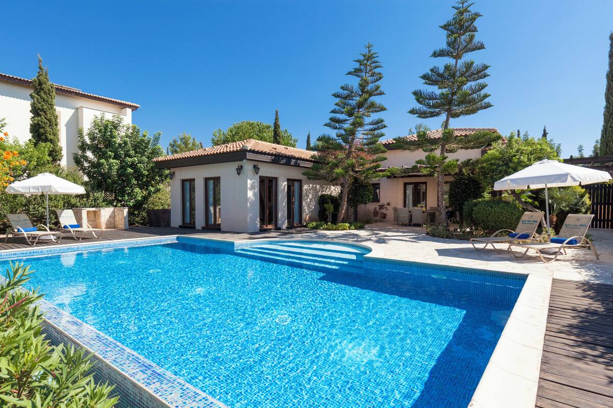 A swimming pool with lounge chairs and umbrellas, surrounded by trees and buildings.