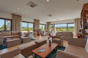 Un salón amplio y luminoso con diversas opciones de asientos, grandes ventanales con vistas al exterior y una estantería llena de libros.
