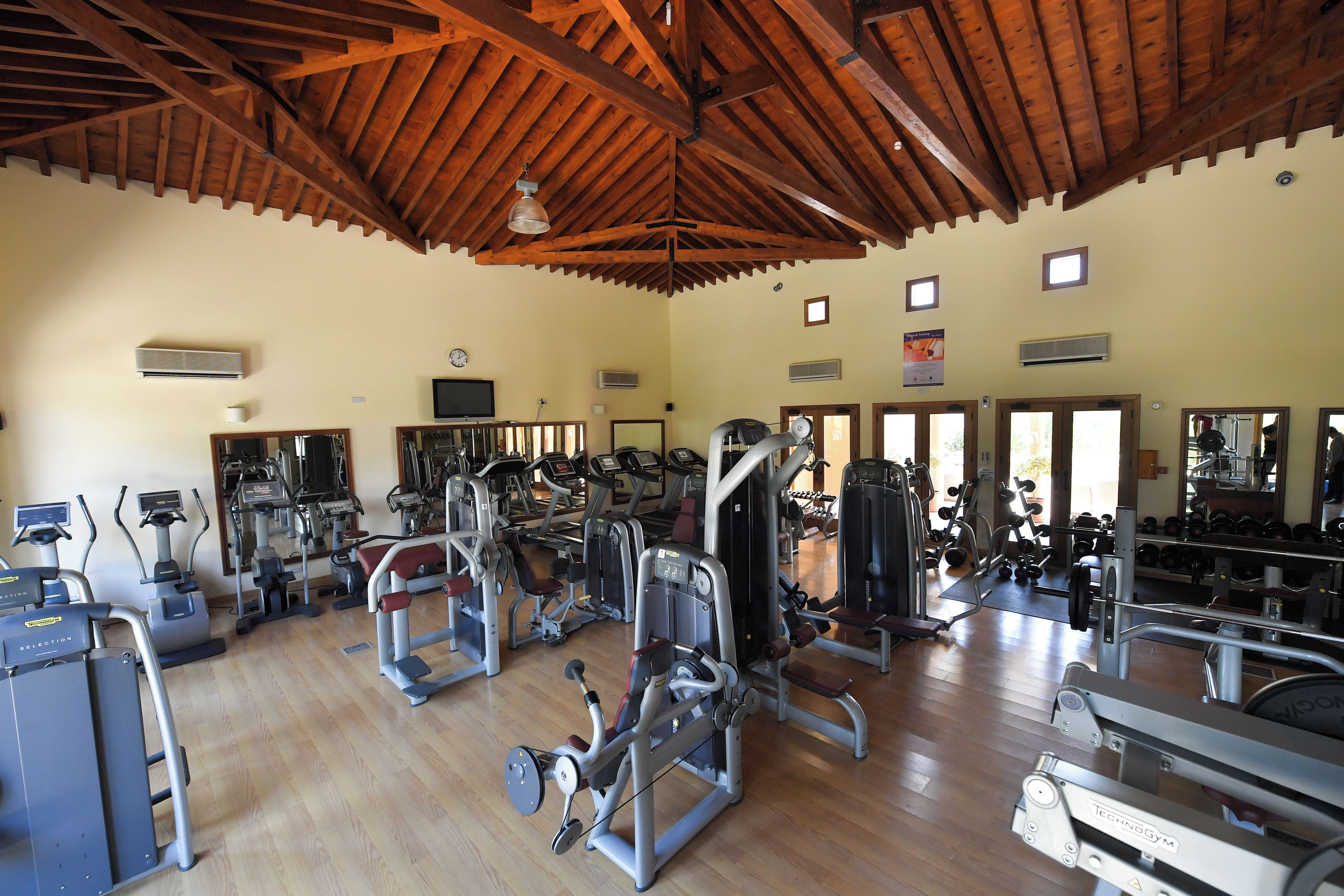Une salle de sport équipée de divers appareils d’exercice, avec des poutres en bois au plafond et une horloge au mur.