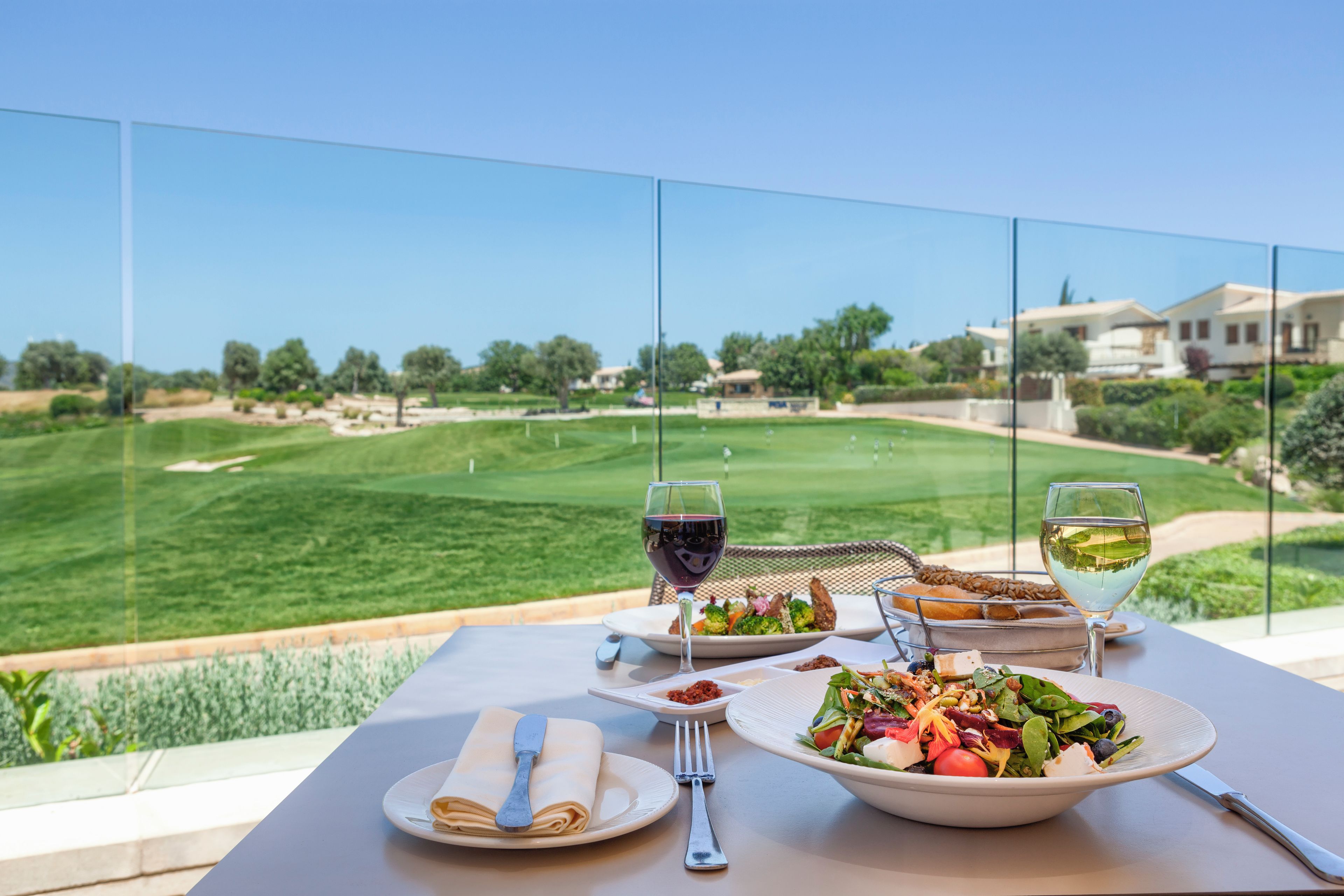 A dining table with plates of food, glasses of wine, and cutlery, overlooking a golf course.