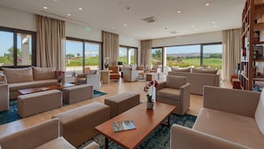 A spacious, well-lit lounge area with multiple seating options, large windows offering a view of the outdoors, and a bookshelf filled with books.