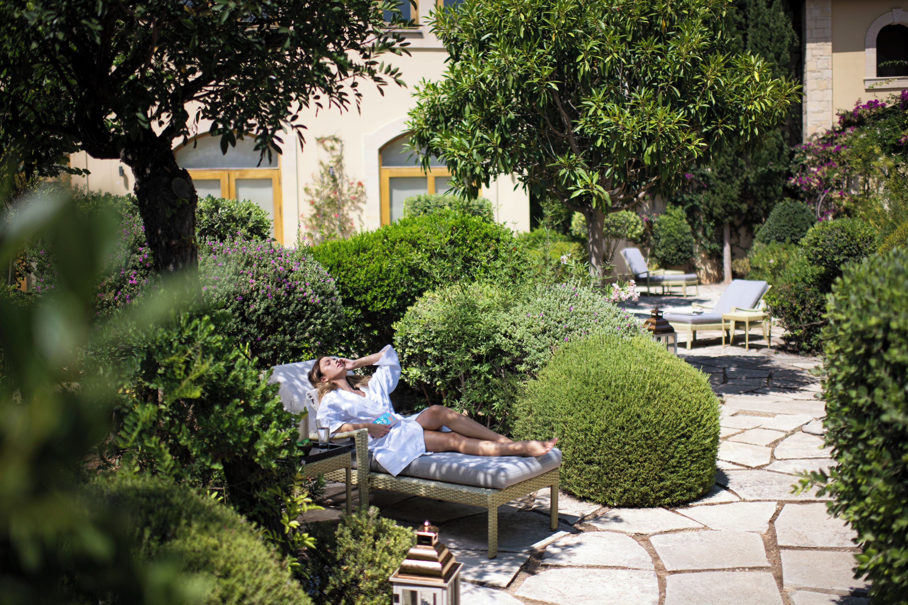 A person relaxing on a wicker lounge chair in a garden with stone pathways, surrounded by lush greenery and a building with a yellow door in the background.