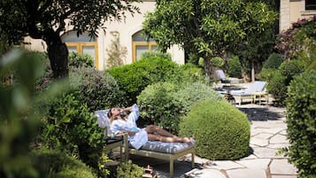 Eine Person entspannt auf einer Korbschaukel in einem Garten mit Steinwegen, umgeben von üppigem Grün und einem Gebäude mit gelber Tür im Hintergrund.