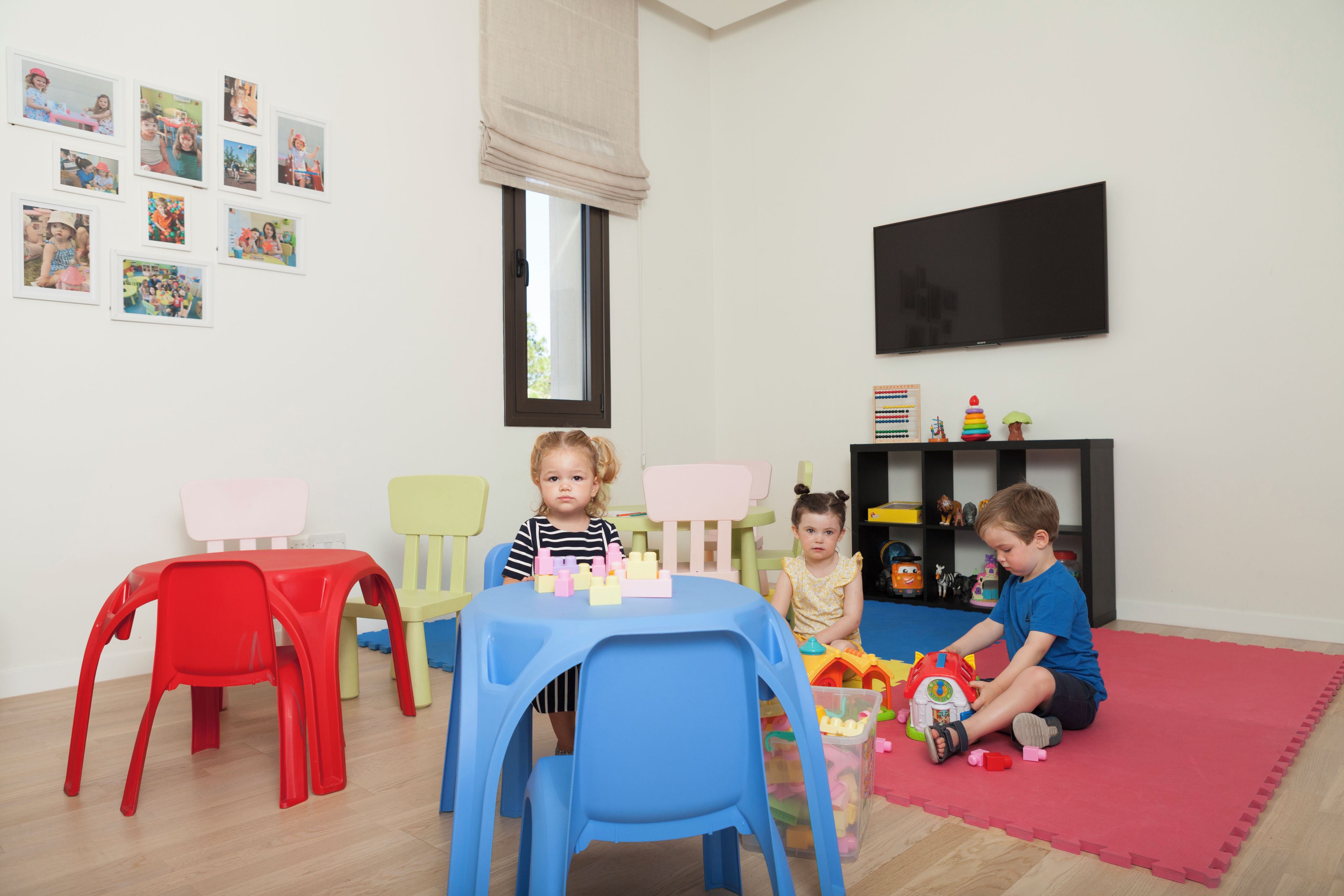 Een kamer met een blauwe tafel, rode en gele stoelen, een televisie en een boekenkast met speelgoed.