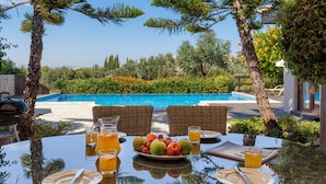 A dining table with fruit, drinks, and a view of a swimming pool and greenery.