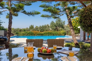 A dining table with fruit, drinks, and a view of a swimming pool and greenery.