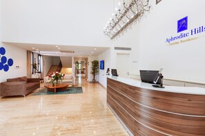 Un área de recepción con un mostrador de madera, una araña de cristal y un cartel que indica 'Aphrodite Hills Holiday Residences'.