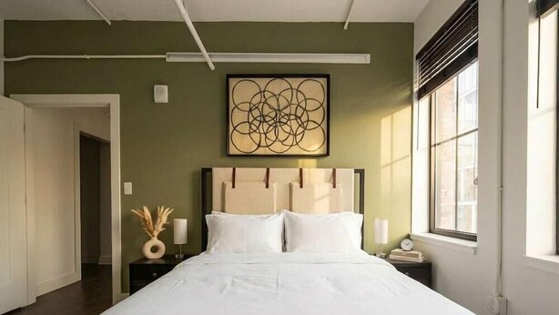 A neatly made bed in a room with a green wall, a large abstract painting, and a window with blinds. - Boulevard Stays Downtown Nashville (Nashville)