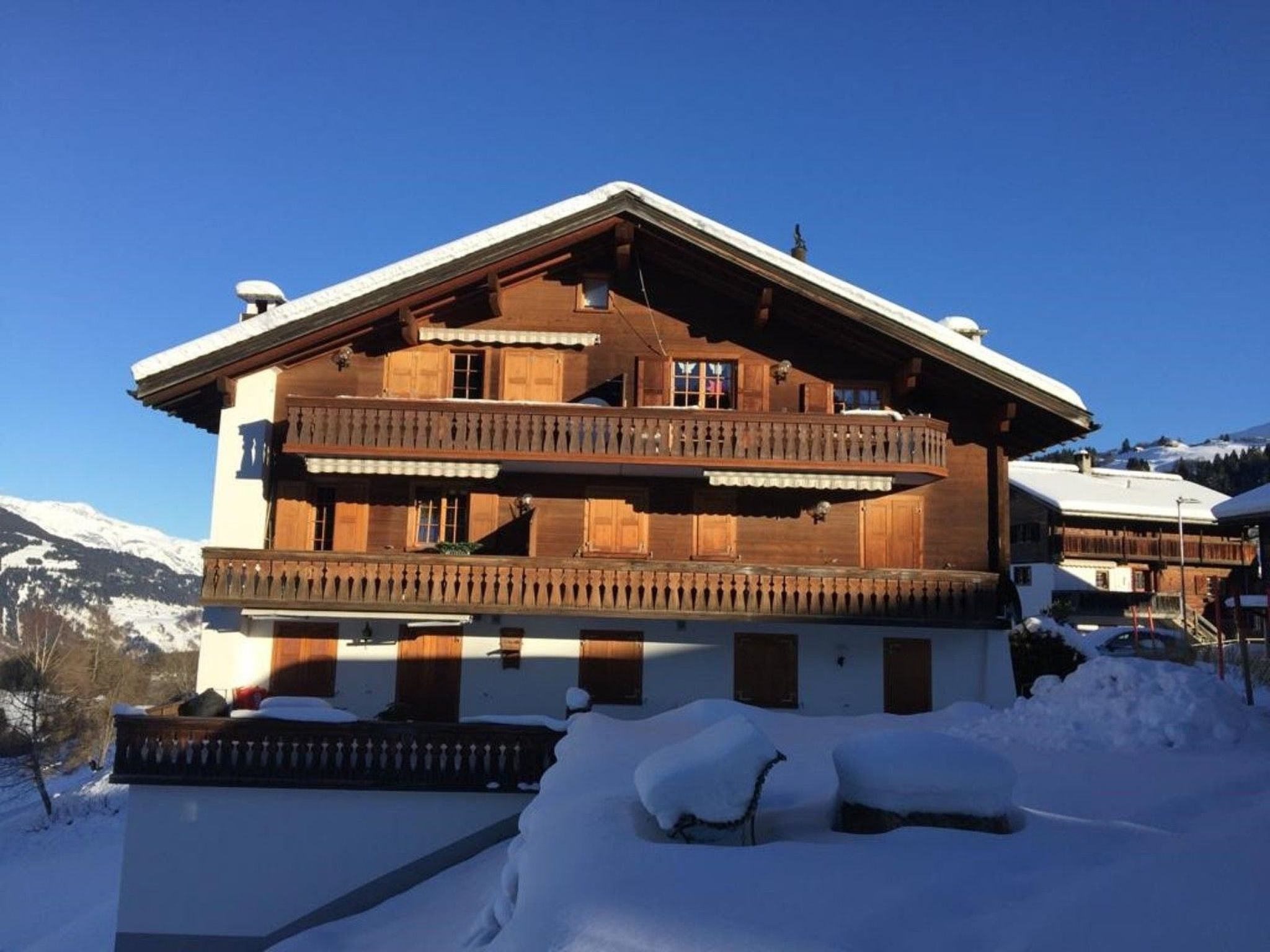 A two-story wooden chalet with a balcony, surrounded by snow.