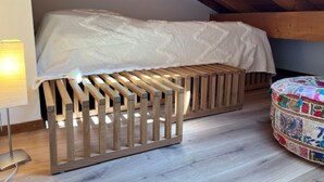 Ein Schlafzimmer im Loft-Stil mit schräg verlaufender Decke, einem Holzbettgestell und einem farbenfrohen Hocker.