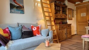 Ein gemütlicher Wohnbereich mit einer Couch, bunten Kissen, einem kleinen Tisch mit Kerze, einer Holzleiter und einer Küchenzeile mit Holzsäulen.