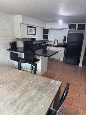 Une cuisine avec des meubles blancs, un réfrigérateur noir et un coin repas comprenant une table et des chaises en bois.