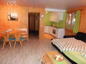 A room with a bed, a desk with chairs, a kitchen area, and a door leading to another room. - Schweizer, Obersaxen-Affeier, (Obersaxen), 34006B by Interhome (Obersaxen Affeier)