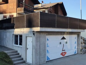 A building with a garage door featuring a face design, a white door, and a set of stairs leading to a balcony. - Schweizer, Obersaxen-Affeier, (Obersaxen), 34006B by Interhome (Obersaxen Affeier)