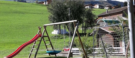 Un terrain de jeux avec un toboggan et des balançoires, entouré d’un champ d’herbe et avec des maisons en arrière-plan.