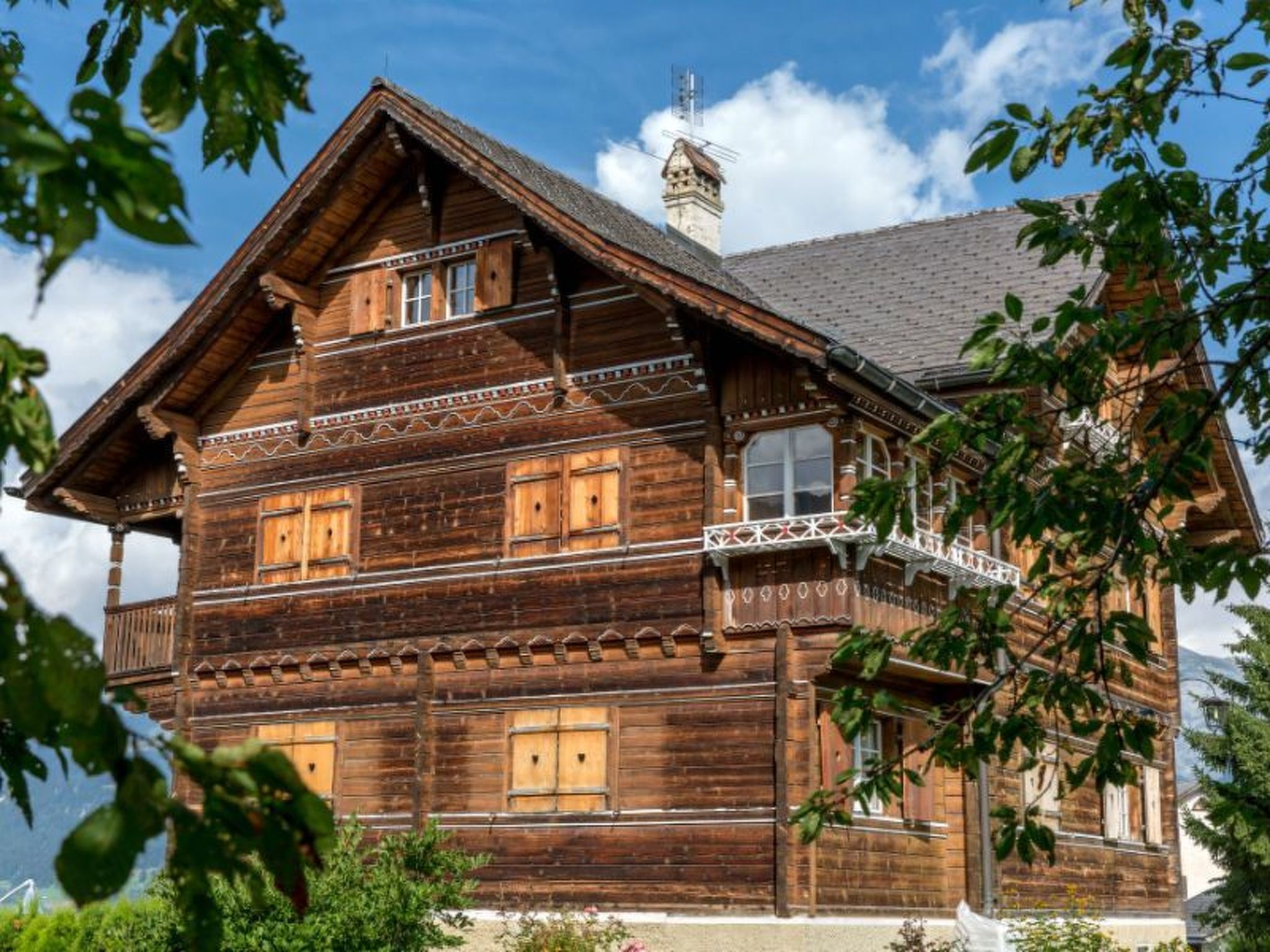 Une maison en bois à plusieurs étages, dotée d’un balcon, d’une cheminée et d’une fenêtre ornée d’un motif décoratif.