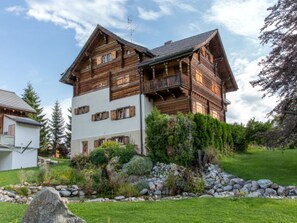 Un chalet en bois à plusieurs étages, doté de balcons, entouré d’un jardin bien entretenu et d’un petit étang.