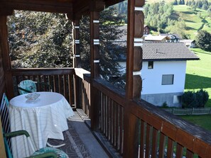 A wooden balcony with a table and chairs, overlooking a green landscape and a white building. - Gyrgarta, Obersaxen-Meierhof, (Obersaxen), 34066B by Interhome (Obersaxen Affeier)