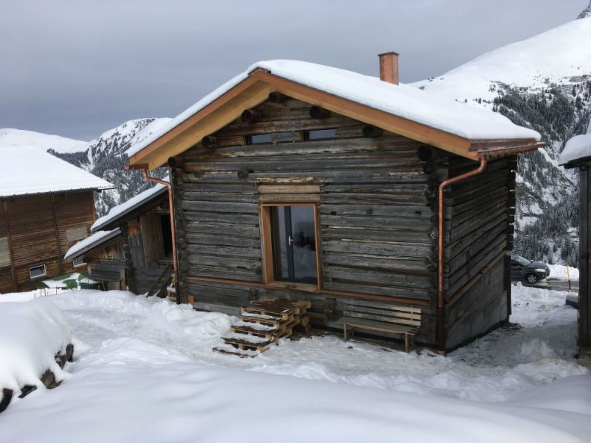 Une cabane en bois, dotée d’un toit en pente et d’une cheminée, entourée de montagnes enneigées.
