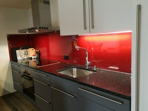 Cocina moderna con pileta de acero inoxidable, encimera de granito negro y salpicadero rojo.