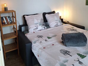 Un dormitorio con cama, almohadas, una mesita de noche con libros y una lámpara.