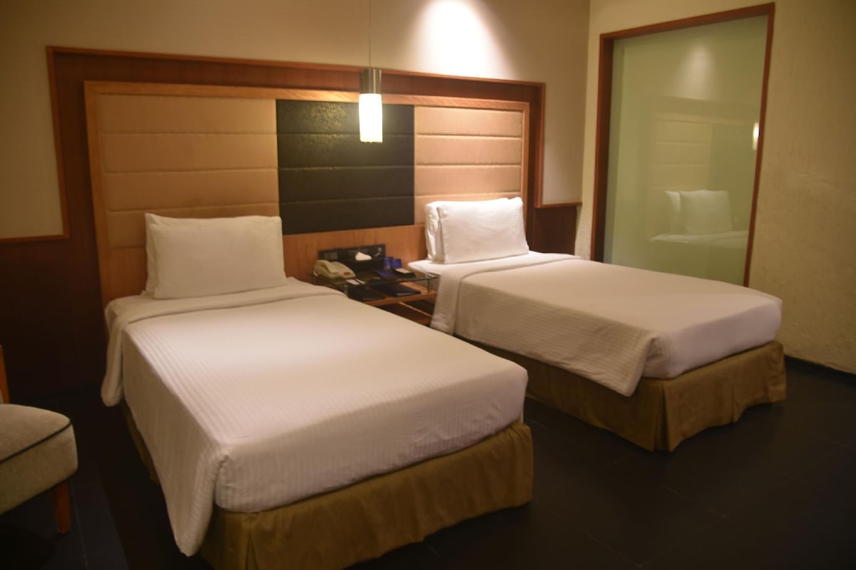 Habitación de hotel con dos camas individuales, cabecero de madera, mesita de noche con teléfono y puerta de vidrio que da a un baño.