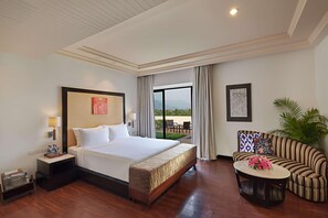 Quarto de hotel com uma cama grande, um sofá, uma pequena mesa com flores, uma mesinha de cabeceira e vista para o exterior.