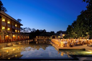 Uma área à beira da piscina com mesas e cadeiras, um edifício com vários andares e árvores ao fundo.