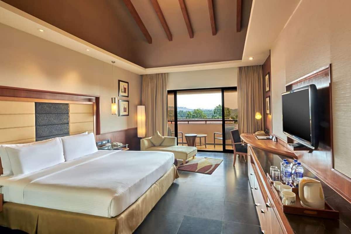 A hotel room with a large bed, a desk with a television, a balcony with a view, and a dining area.
