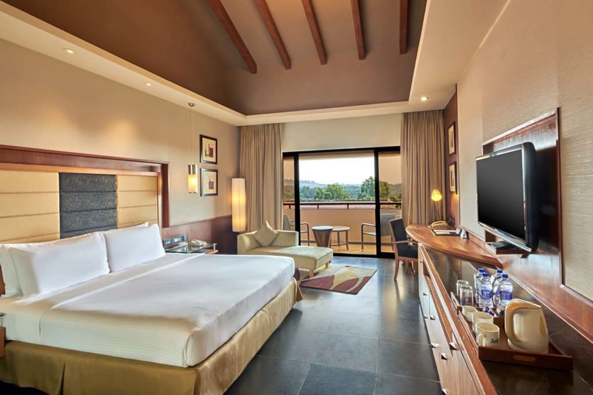 Habitación de hotel con una cama grande, un escritorio con televisor, un balcón con vistas y un comedor.