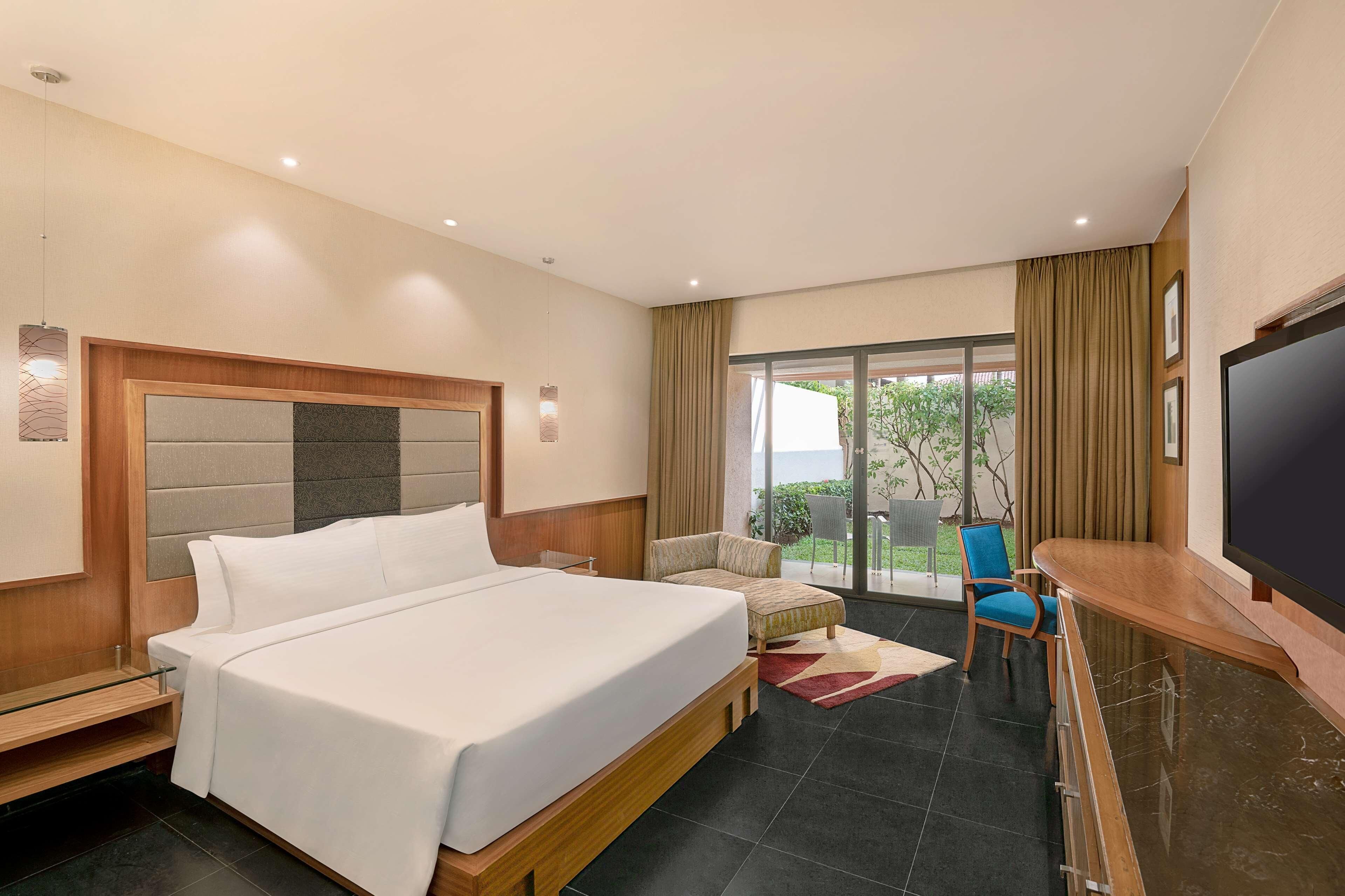 Una habitación de hotel moderna con una cama grande, televisor, zona de estar con una silla y vistas al exterior a través de un ventanal.