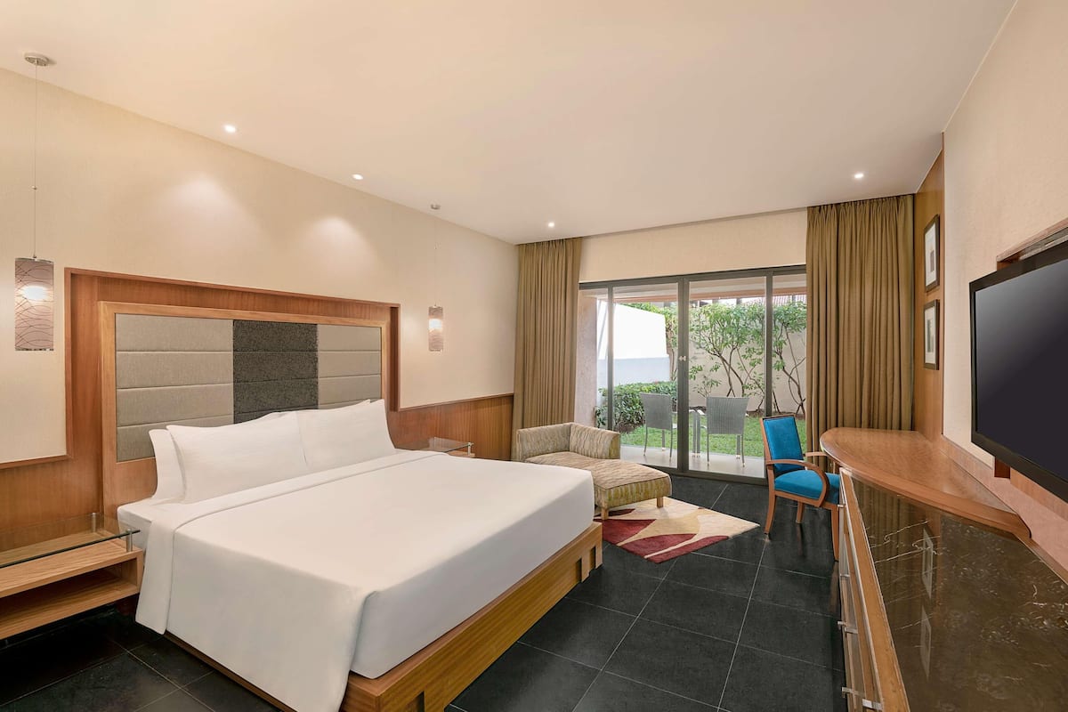 A modern hotel room with a large bed, a TV, a seating area with a chair, and a view of the outdoors through a glass door.