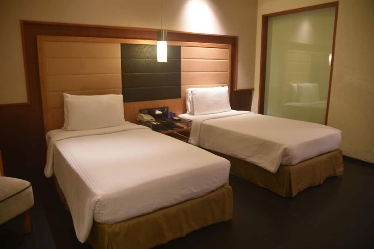Habitación de hotel con dos camas individuales, cabecero de madera, mesita de noche con teléfono y puerta de vidrio que da a un baño.