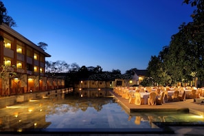 A poolside area with tables and chairs, a building with multiple floors, and trees in the background. - Radisson Blu Resort & Spa Alibaug (Alibag)