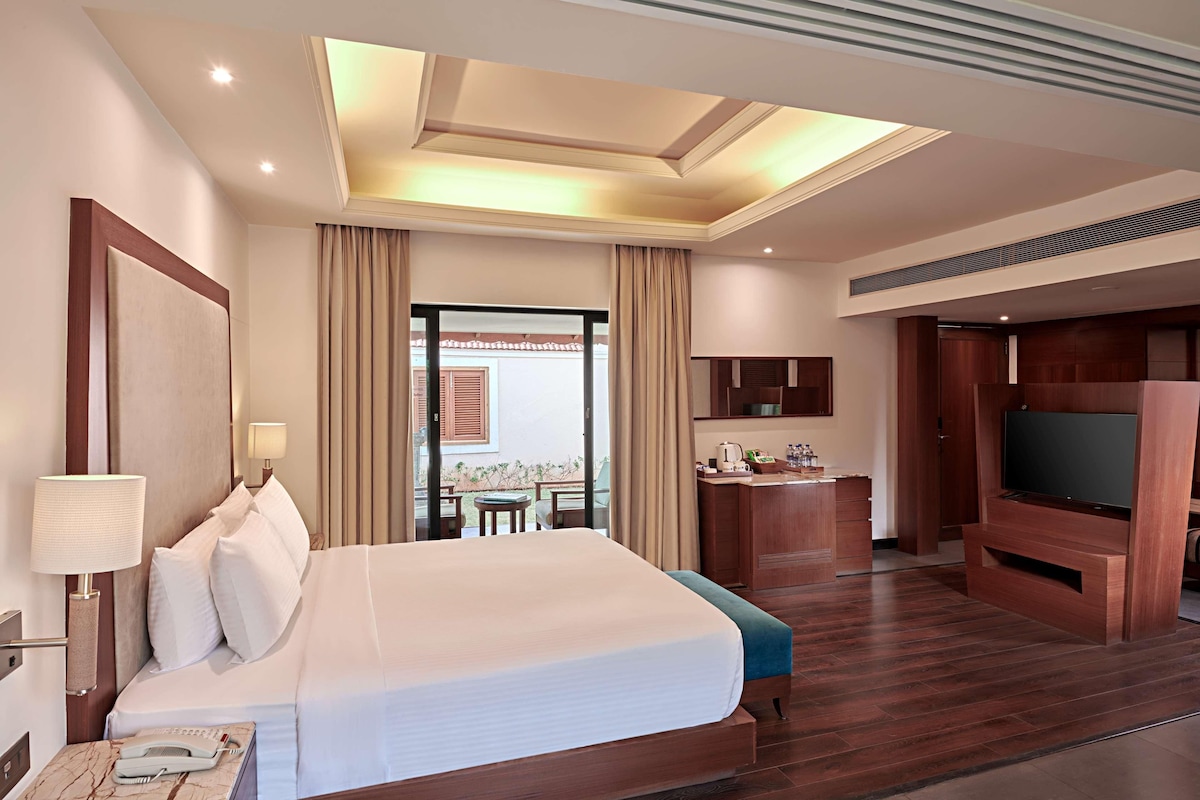 Habitación de hotel con una cama grande, televisión, escritorio con silla y balcón con cortinas.