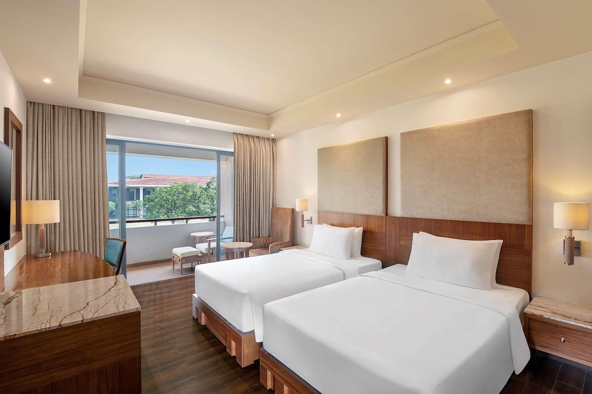 A hotel room with two beds, a desk with a lamp, a chair, a small table, and a view of the outside.