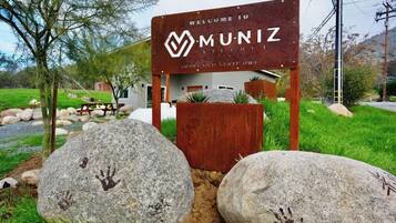 Un cartello di benvenuto per Muniz Village con una struttura in legno rustica, grandi rocce con impronte di mani e un paesaggio erboso.
