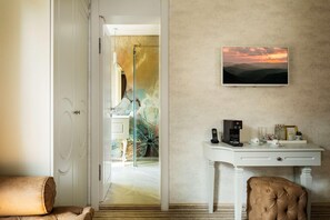 A hotel room with a white desk, a television mounted on the wall, and a view of a sunset through the window.