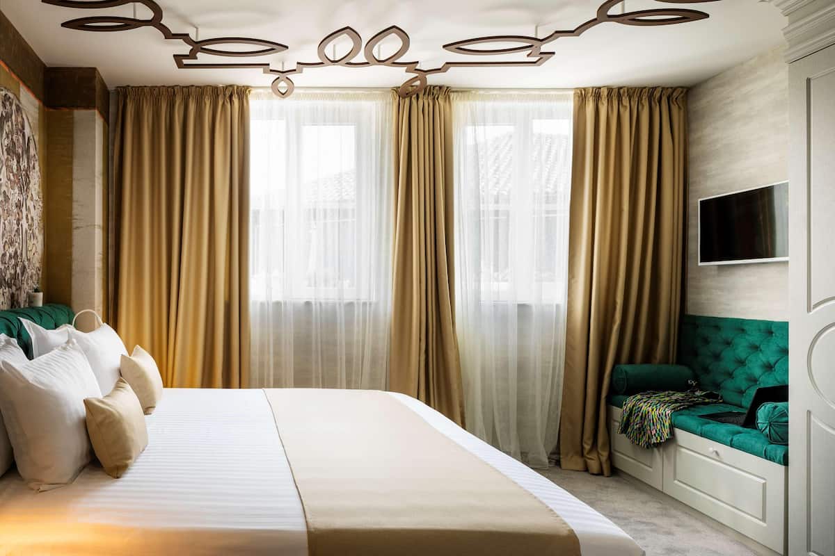 A hotel room with a large bed, beige curtains, a green upholstered bench, and a flat-screen TV mounted on the wall.