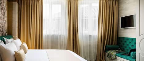 Una habitación de hotel con una cama grande, cortinas color beige, un banco tapizado en verde y un televisor de pantalla plana montado en la pared.